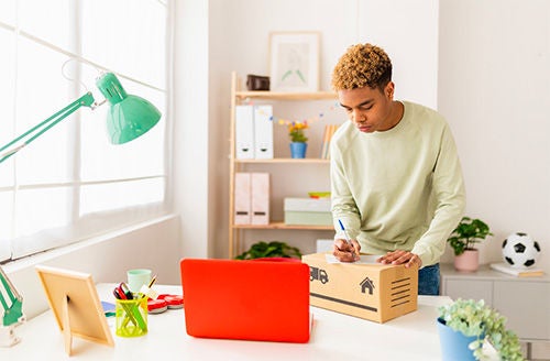 A young man stands next to a desk and writes on top of a cardboard box while looking at a laptop in front of him.