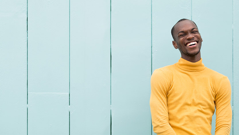 A man smiles at the camera while sitting against a wall.