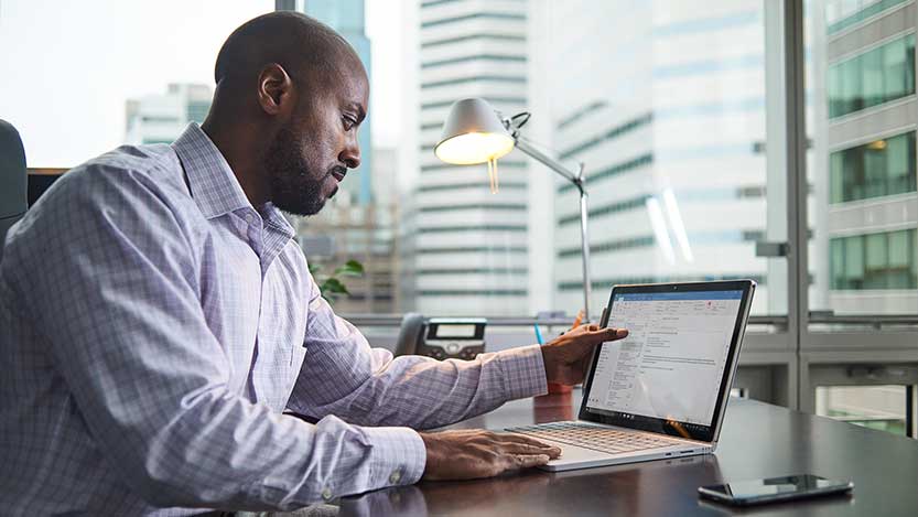 A man sits at a desk in an office with large windows and works on a laptop computer.