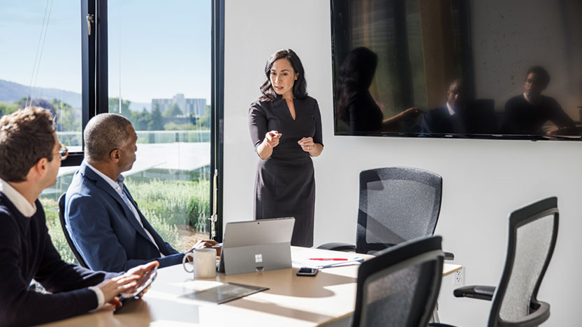 In an office, a woman stands in front of two men sitting at a table and explains something while pointing at a laptop.