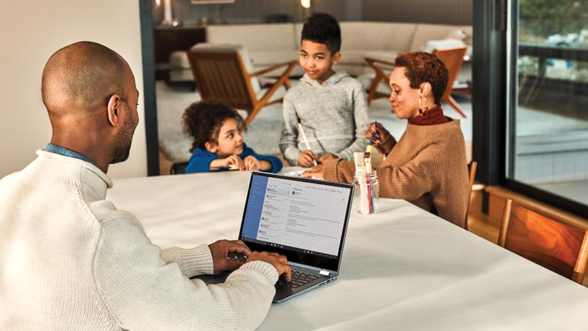 A man works on his computer while a woman and two children sit across a table.