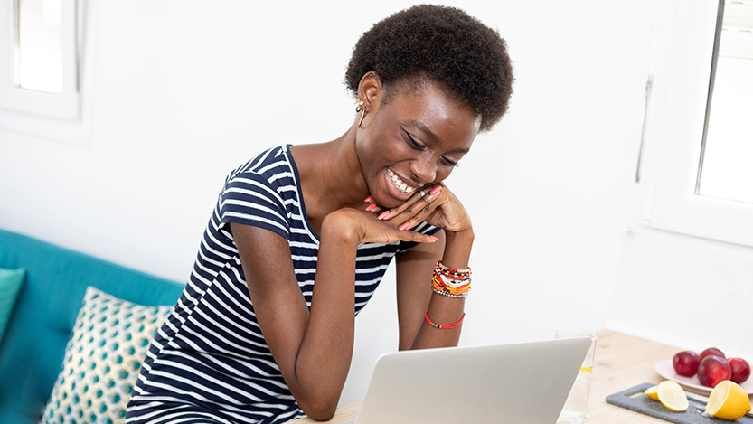 A woman sitting at a table smiles while looking at a laptop computer. 