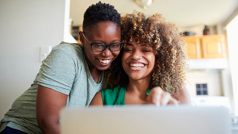 Two women hug and smile while looking at a laptop computer.