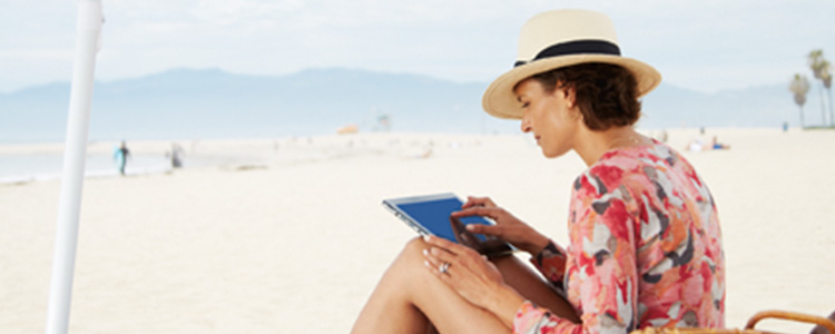 Une femme avec un chapeau, assise sur la plage, utilise une tablette.